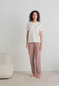 T-shirt crème à manches courtes associé à un pantalon large rose clair, tous deux en tissu doux, se tenant sur une surface en moquette claire.