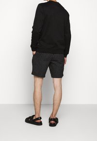 Sudadera negra de mangas largas, combinada con pantalones cortos a rayas en gris y blanco. Sandalias oscuras complementan el conjunto, con un fondo blanco liso.