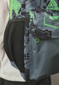 Mochila con un exterior de tela gris, acentos verdes y un bolsillo lateral de malla. Presenta patrones abstractos y un pequeño detalle en forma de lazo verde.
