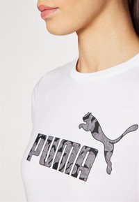 White cotton t-shirt featuring a large, gray camouflage Puma logo and text. Rounded neckline and short sleeves.