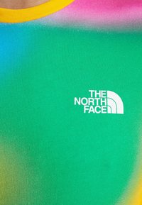 Detailní záběr na vícebarevnou košili se žlutým výstřihem a logem The North Face v bílé barvě na zelené látce.