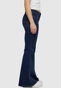 Donkerblauwe denim flared jeans met een middenhoge taille, vijf zakken en stikselaccenten, gecombineerd met witte sneakers.
