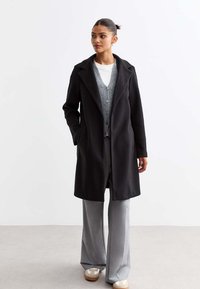 Manteau en mélange de laine noire avec un col cranté, fermeture à un bouton, poches latérales, porté sur un cardigan gris et un pantalon gris clair.
