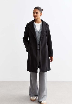Black wool blend coat with a notched collar, single button closure, side pockets, worn over a gray cardigan and light gray trousers.