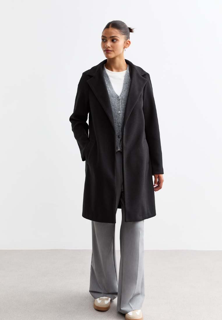 Manteau en mélange de laine noire avec un col cranté, fermeture à un bouton, poches latérales, porté sur un cardigan gris et un pantalon gris clair.