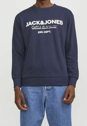Person trägt einen marineblauen Pullover mit weißem "JACK & JONES" Textlogo und blaue Jeans, steht vor einem einfarbigen Hintergrund.