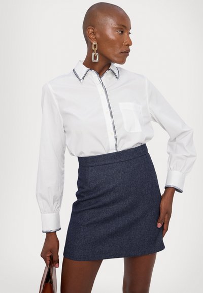 White collared shirt with navy stitching and a pocket, paired with a fitted dark blue skirt. Model holds a clutch bag.