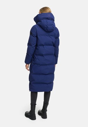 Lange donsjas in donkerblauw, met een hoge kraag, capuchon en gestikt ontwerp. Inclusief zijzakken en gedragen over donkere broek en laarzen.