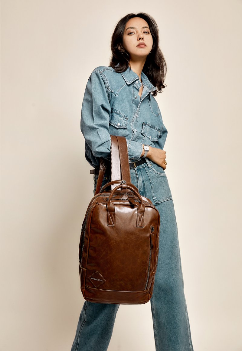 Smeđa kožna naprtnjača s teksturiranom završnom obradom, dvostrukim ručkama na vrhu, podesivim remenom za rame i prednjim zip džepom. Model nosi traper odijelo.