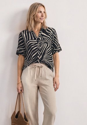 Kvinde står, iført en sort og hvid zebrastribet skjorte, beige bukser med snøre, holder en fletnettaske, kigger til siden.