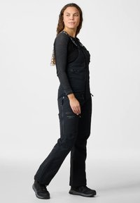 Donna che indossa pantaloni da esterno neri con bretelle e camicia a maniche lunghe, in piedi e che guarda di lato su uno sfondo grigio uniforme.