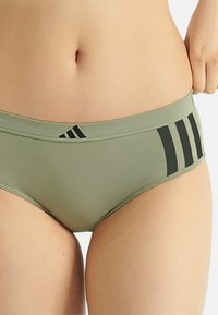 Caleçon vert olive avec une texture lisse, arborant un logo noir et trois bandes horizontales sur le côté. Design minimaliste avec une coupe confortable.