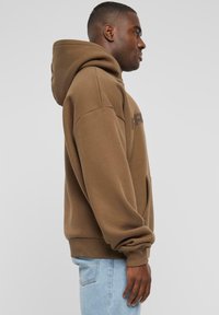 Brauner Kapuzenpullover aus weichem Material, mit einer Fronttasche und gerippten Bündchen. Einfaches Design mit Logodetail auf der Brust.