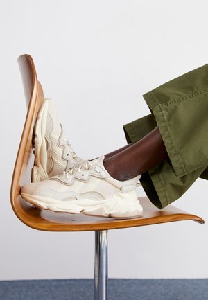 Fødder iført beige sneakers, der hviler på en træstol, med ben iført grønne bukser mod en ensfarvet hvid baggrund.