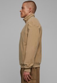 Beige fleece genser med høy krage, glidelås og en frontlomme. Har ribbestrikkede mansjetter og hem. Glatt tekstur og avslappet passform.