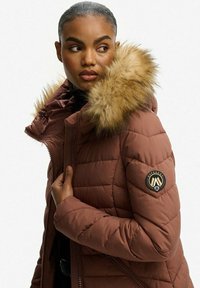 Brązowa kurtka puchowa z kapturem z sztucznego futra, pikowane wykończenie, zapinana na zamek z przodu i z bocznymi kieszeniami. Posiada naszywkę z logo na rękawie.