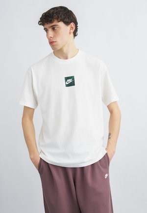 Ung mand med mørkt krøllet hår iført en hvid Nike T-shirt og mørkerøde Nike joggingbukser, stående med hænderne i lommerne og ser mod venstre.