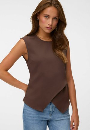 Jeune femme aux longs cheveux bruns porte un haut marron sans manches avec un ourlet asymétrique et un jean bleu clair sur un fond uni.