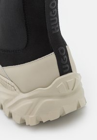Zapatilla alta en negro y beige con suela de goma texturizada, diseño elegante y distintiva marca "HUGO" en el lateral.