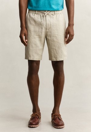 Mann trägt beige Shorts mit Kordelzug, ein türkisfarbenes Hemd und braune Leder-Schnürschuhe, steht vor einem schlichten Hintergrund.