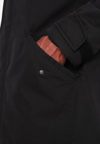 Eine dunkelhäutige Hand steckt in der Seitentasche einer schwarzen Jacke mit Druckknopf und Klettverschluss am Ärmel.