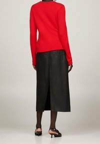 Roter Wollpullover mit langen Ärmeln, kombiniert mit einem schwarzen Leder-Midrock mit einem Schlitz hinten und zwei hinteren Taschen. Schwarze Absatzmules.