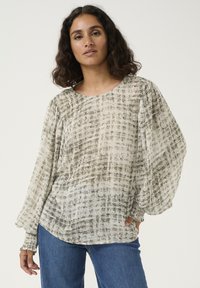 Blouse transparente à manches longues en beige clair avec un motif à carreaux subtil, coupe décontractée et poignets froncés. Associée à un jean en denim bleu.