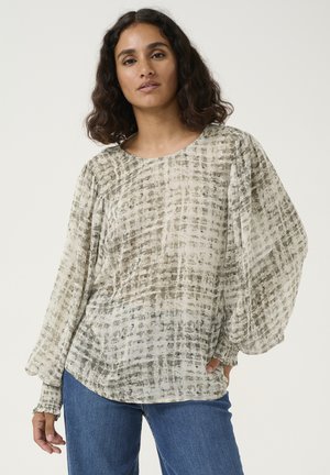 Sheer, long-sleeve blouse in light beige with a subtle grid pattern, relaxed fit, and gathered cuffs. Paired with blue denim jeans.