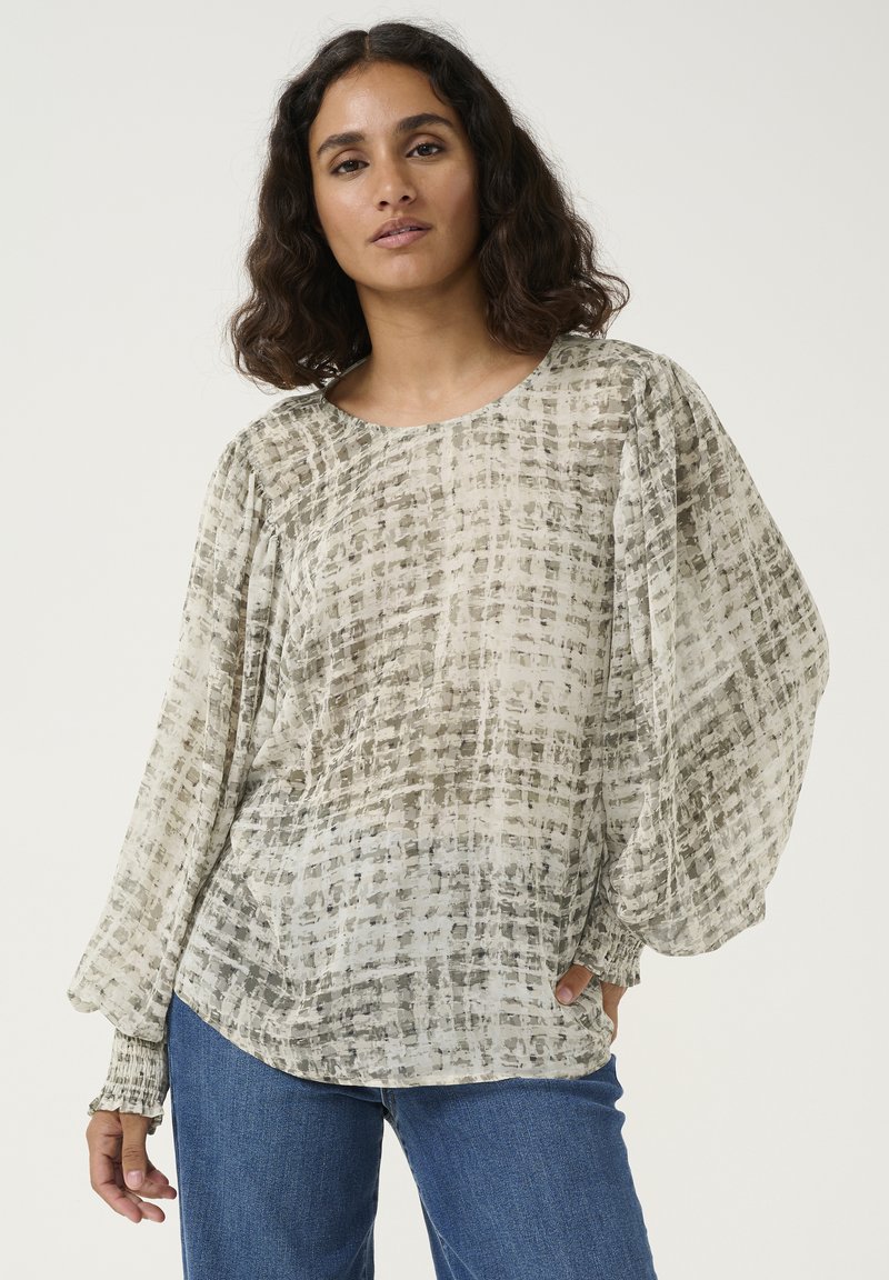 Blouse transparente à manches longues en beige clair avec un motif à carreaux subtil, coupe décontractée et poignets froncés. Associée à un jean en denim bleu.