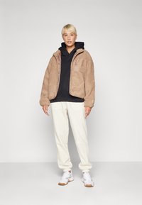 Beige fluffy jakke over en sort hoodie, parret med hvide sweatpants og hvide sneakers. Jakken har lynlås og frontlomme.