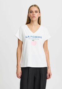 Balta medvilninė marškinė su V formos kaklu ir trumpomis rankovėmis. Priekyje yra mėlyni užrašai "LA POMME" ir rožinė obuolio grafika, suteikianti kasdienio stiliaus.