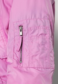 Närbild av en rosa jackärm med en silverfärgad dragkedjeficka och sydd rektangulär detalj på en slät tyg.