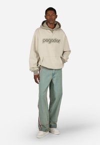 Ανοιχτό καφέ φούτερ με κείμενο "pagador" σε σκουρό καφέ, συνδυασμένο με φαρδιά ανοιχτό γαλάζια τζιν παντελόνια με πλαϊνές ρίγες και λευκά αθλητικά παπούτσια.