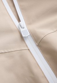 Tissu beige avec une fermeture éclair en plastique blanc dotée d'une languette texturée, présenté en gros plan.