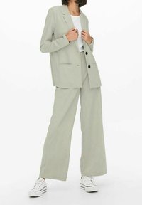 Lichtgrijze blazer met twee voorzakken, gecombineerd met een broek met wijde pijpen en een wit shirt. Schoeisel: witte sneakers met zwarte strepen.