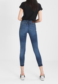 Tunna blå denimjeans med hög midja, fransad kant och subtila slitningar, i kombination med en svart kortärmad topp och sandaler med klack och remmar.