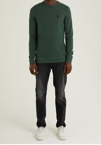 Dunkelgrüner Pullover mit strukturiertem Strickmuster, runder Ausschnitt und kleinem Logopatch auf der Brust. Kombiniert mit dunklen Jeans und weißen Sneakern.