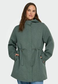 Zelena kapuca jakna s vrvico na pasu, narejena iz mehke tkanine, z dvema sprednjima žepoma in sproščenim krojem.