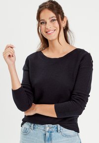 Black textured sweater with a wide neckline, long sleeves, and metal accents at the shoulder, paired with light blue jeans.
