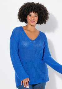 Maglione blu lavorato a maglia con scollatura a V, vestibilità ampia e maniche lunghe. Presenta un design testurizzato e orlo a coste. Indossato con jeans in denim.