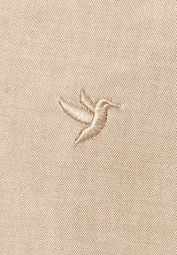 Tejido beige que presenta un delicado colibrí bordado en un tono ligeramente más claro, mostrando un intrincado trabajo de costura y alas detalladas.