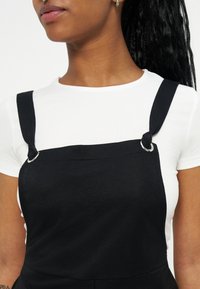 Γυναίκα που φοράει λευκό κοντομάνικο πουκάμισο κάτω από μαύρο αμάνικο φόρεμα με δεμένους ιμάντες και ασημένιες λεπτομέρειες σε μορφή δακτυλίων.