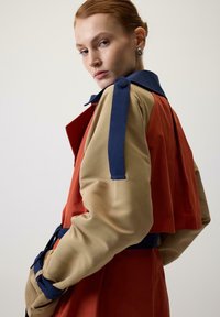 Abrigo con bloques de color que presenta un cuerpo en rojo óxido, mangas beige y acentos en azul marino. Incluye un amplio cuello y una cintura ceñida con cinturón. Textura de tela suave.