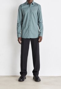 Camicia turchese con bottoni, collo, maniche lunghe e taglio aderente; abbinata a pantaloni neri e sneakers nere.