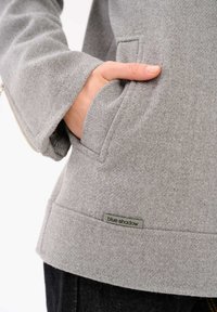 Szara polarowa kurtka z kieszenią, zaprezentowana z ręką opartą na niej. Tkanina jest teksturowana; na dolnym brzegu znajduje się metka "blue shadow".