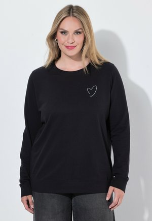 Kobieta o blond włosach, ubrana w czarną koszulkę z długim rękawem, z małym białym serduszkiem po lewej górnej stronie.