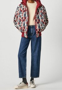 Fyllig jacka med röd huva och ett mönster i hela plagget i kräm, marinblått och rött. Kombinerad med blå vida jeans och vita sneakers.