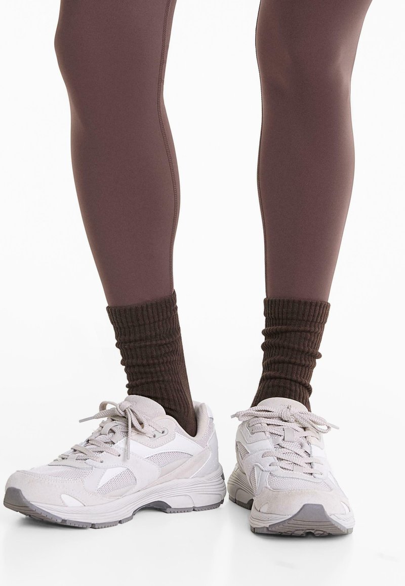 Benen in bruine leggings, donkerbruine gestrikte sokken en witte chunky sneakers met veters op een witte achtergrond.