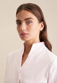 Blusa rosa chiaro con bottoni e colletto rialzato, tessuto liscio e leggero lucido. Design aderente, mostrata su uno sfondo neutro.