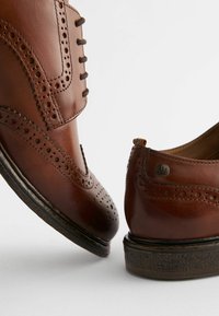Chaussures en cuir marron avec des détails de brogue, présentant un bout arrondi et une semelle sombre contrastée. Comprend des coutures décoratives et un accent en métal.